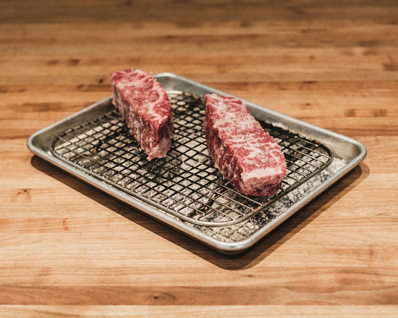 Two pieces of raw Wagyu beef on a metal tray over a wooden counter, ready for cooking.